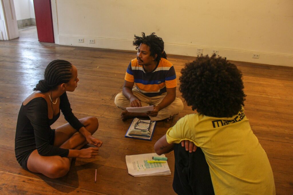 Macacada Três integrantes da Cia de Teatro Negro Macacada sentados no chão em roda, durante um ensaio teatral.
