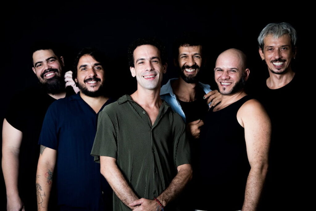 Magiluth Seis integrantes do grupo Magiluth posam sorrindo em estúdio com fundo preto.